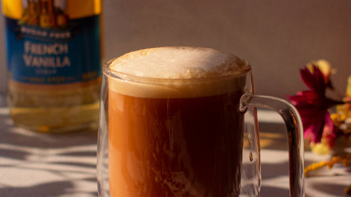 A London Fog in a glass mug in front of florals and a French Vanilla syrup bottle.