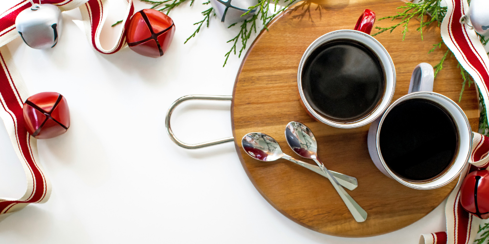 Two cups of coffee on a wood plate, surrounded by red bells and fir tree branches
