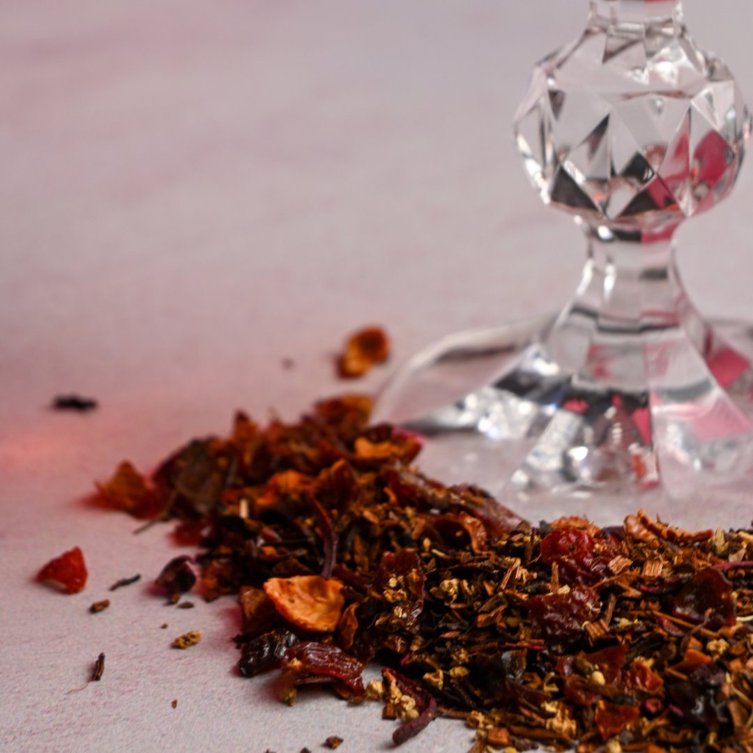Raspberry Rosé organic herbal tea blend, featuring rosehips and hibiscus scattered on tabletop with glass base in view.